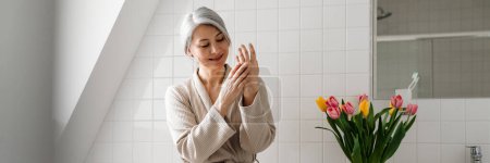 Foto de Mujer sonriente madura aplicando crema mientras está sentada en el baño en casa - Imagen libre de derechos