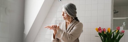 Foto de Mujer gris madura aplicando crema mientras está sentada en el baño en casa - Imagen libre de derechos