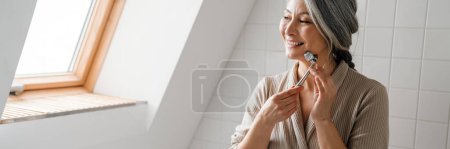 Foto de Mujer gris madura sonriendo mientras masajea la cara con rodillo de jade en el baño en casa - Imagen libre de derechos