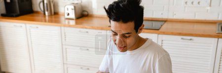 Photo for Young smiling hispanic man reading a book while having breakfast in the kitchen at home, sitting at the table - Royalty Free Image