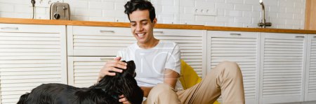 Photo for Smiling hispanic young man petting his dog while sitting on a kitchen floor holding digital tablet computer - Royalty Free Image