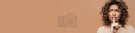 Téléchargez les photos : Jeune femme hispanique fronçant les sourcils tout en montrant un geste de silence à la caméra isolé sur fond beige - en image libre de droit