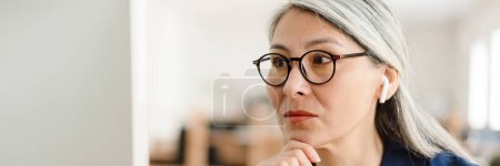 Téléchargez les photos : Le portrait d'une femme asiatique en lunettes et écouteurs regardant l'écran de l'ordinateur assis à la table dans le bureau - en image libre de droit