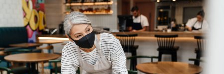 Photo for Mature waitress woman wearing face mask cleaning table in cafe indoors - Royalty Free Image