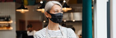 Photo for Mature waitress woman wearing face mask standing in cafe indoors - Royalty Free Image