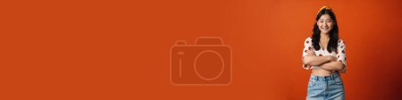 Téléchargez les photos : Souriant jeune femme asiatique en vêtements d'été debout sur fond orange, les bras croisés, regardant la caméra - en image libre de droit