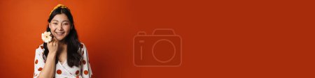Téléchargez les photos : Souriant jeune femme asiatique en vêtements d'été debout sur fond orange, tenant fleur - en image libre de droit