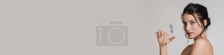 Téléchargez les photos : Jeune femme blanche aux cheveux bruns tirée vers le haut tenant friseur de cils isolé sur fond gris - en image libre de droit