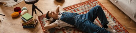 Téléchargez les photos : Jeune homme blanc dans les écouteurs se reposant tout en travaillant sur un tapis artisanal à la maison - en image libre de droit