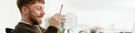 Téléchargez les photos : Jeune homme roux gesticuler et travailler avec un ordinateur portable tout en étant assis à la table dans le bureau - en image libre de droit