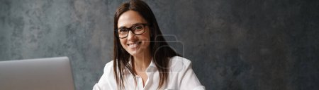 Photo for Smiling mid aged brunette businesswoman in shirt sitting at the desk with laptop computer over dark background, holding mobile phone - Royalty Free Image