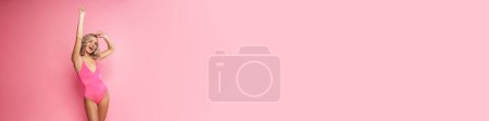 Foto de Retrato de una joven rubia sonriente con traje de baño de pie sobre el fondo de la pared de color rosa, manos desdichadas - Imagen libre de derechos