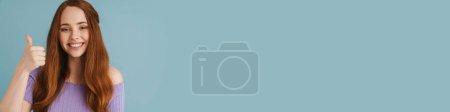 Téléchargez les photos : Jeune femme rousse souriante et montrant pouce vers le haut à la caméra isolé sur fond bleu - en image libre de droit