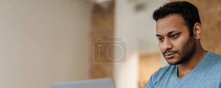 Foto de Oriente Medio hombre sin afeitar trabajando con el ordenador portátil mientras está sentado en el escritorio en la oficina - Imagen libre de derechos