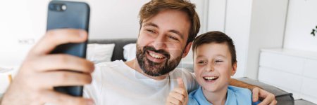 Téléchargez les photos : Père heureux et petit fils prenant selfie via smartphone assis sur le canapé à la maison - en image libre de droit