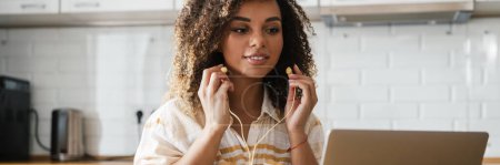 Foto de Una mujer negra confiada que se pone auriculares con cable y mira la pantalla del portátil mientras está sentada en la cocina ligera - Imagen libre de derechos