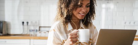 Téléchargez les photos : Une femme noire souriante dans un casque filaire avec une tasse dans ses mains assis devant un ordinateur portable dans la cuisine blanche - en image libre de droit