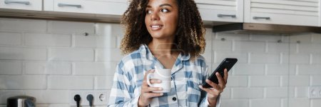 Téléchargez les photos : Le portrait d'une femme noire souriante en pyjama regardant sur le côté avec une tasse et un téléphone dans ses mains tout en se tenant dans la cuisine blanche - en image libre de droit