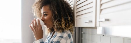 Téléchargez les photos : Portrait d'une femme noire en pyjama à carreaux buvant dans la tasse assise sur le comptoir de la cuisine devant la fenêtre - en image libre de droit