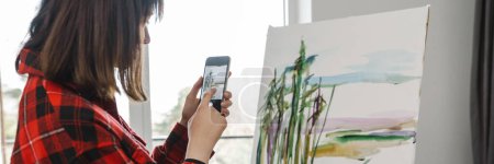 Téléchargez les photos : Jeune peintre blanche tenant un pinceau debout à l'intérieur de la toile en prenant des photos de la toile - en image libre de droit
