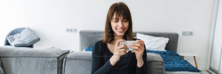 Téléchargez les photos : Jeune femme souriante utilisant un téléphone portable alors qu'elle était assise sur un canapé à la maison - en image libre de droit