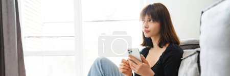 Téléchargez les photos : Souriant jeune femme tenant téléphone portable textos tout en étant assis appuyé sur un canapé à la maison - en image libre de droit