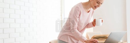 Téléchargez les photos : Joyeux jeune femme blanche portant des vêtements de salon debout à la table avec ordinateur portable, étude, appel vidéo, tenant tasse - en image libre de droit