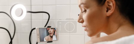 Téléchargez les photos : Jeune femme blanche souriante blogueuse beauté en bahtrobe faire une vidéo sur téléphone portable dans la salle de bain en appliquant de la crème visage à partir d'un tube arrière - en image libre de droit