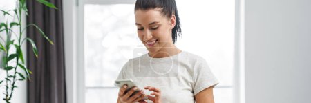 Photo for European smiling woman using mobile phone and making salad in kitchen at home - Royalty Free Image