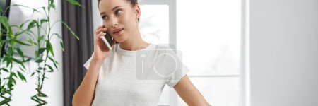 Photo for European young woman talking on mobile phone while having breakfast in kitchen at home - Royalty Free Image
