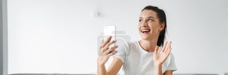 Foto de Mujer sonriente europea haciendo gestos y tomando selfie en el teléfono móvil en casa - Imagen libre de derechos