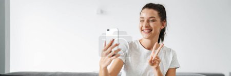 Foto de Mujer sonriente europea haciendo gestos y tomando selfie en el teléfono móvil en casa - Imagen libre de derechos