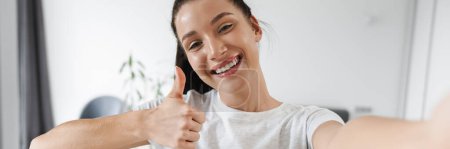Photo for Smiling brunette woman showing thumb up while taking selfie photo at home - Royalty Free Image
