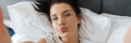 Téléchargez les photos : Brunette femme faire baiser lèvres et de prendre des photos selfie tout allongé sur le lit à la maison - en image libre de droit