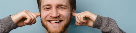 Téléchargez les photos : Barbu gingembre homme souriant tout en bouchant ses oreilles isolées sur fond bleu - en image libre de droit