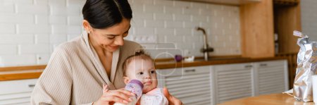 Photo for Young brunette mother smiling and feeding her baby in kitchen at home - Royalty Free Image