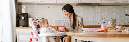Téléchargez les photos : Jeune brune mère souriante et nourrissant son bébé dans la cuisine à la maison - en image libre de droit