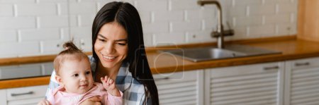 Téléchargez les photos : Jeune mère souriant et tenant son bébé tout en travaillant avec un ordinateur portable dans la cuisine à la maison - en image libre de droit
