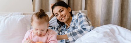 Téléchargez les photos : Jeune mère souriant et jouant avec son bébé tout en étant couché dans le mauvais à la maison - en image libre de droit