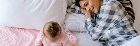 Photo for Young mother smiling and playing with her baby while lying in bad at home - Royalty Free Image