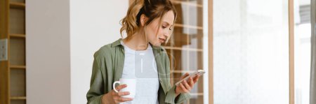 Foto de Mujer blanca joven usando teléfono inteligente mientras bebe café en casa - Imagen libre de derechos