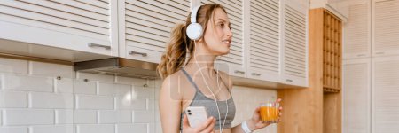 Photo for Smiling young white woman in sportswear drinking juice listening to music with headphone in the kitchen at home - Royalty Free Image