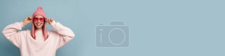 Téléchargez les photos : Jeune femme aux lunettes de soleil roses souriant et regardant la caméra isolée sur fond bleu - en image libre de droit