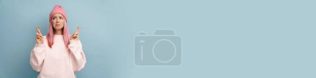 Téléchargez les photos : Jeune femme blanche aux cheveux roses fronçant les sourcils et gardant les doigts croisés isolés sur un mur bleu - en image libre de droit