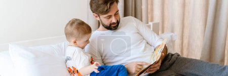 Téléchargez les photos : Père blanc et fils lisant le livre tout en étant couché sur mauvais à la maison - en image libre de droit