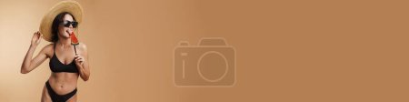 Téléchargez les photos : Jeune femme brune en paille chapeau et lunettes de soleil léchant sucette isolé fond beige - en image libre de droit