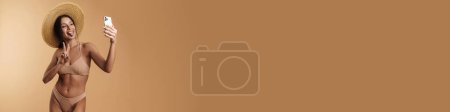 Téléchargez les photos : Jeune femme en maillot de bain montrant sa langue tout en prenant selfie sur téléphone portable isolé sur fond beige - en image libre de droit