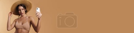 Téléchargez les photos : Jeune femme brune en maillot de bain prenant selfie sur téléphone portable isolé sur fond beige - en image libre de droit