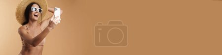 Téléchargez les photos : Jeune femme blanche en maillot de bain souriant et utilisant un téléphone portable isolé sur fond beige - en image libre de droit