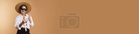 Téléchargez les photos : Jeune femme gestuelle tout en prenant selfie sur téléphone cellulaire isolé sur fond beige - en image libre de droit